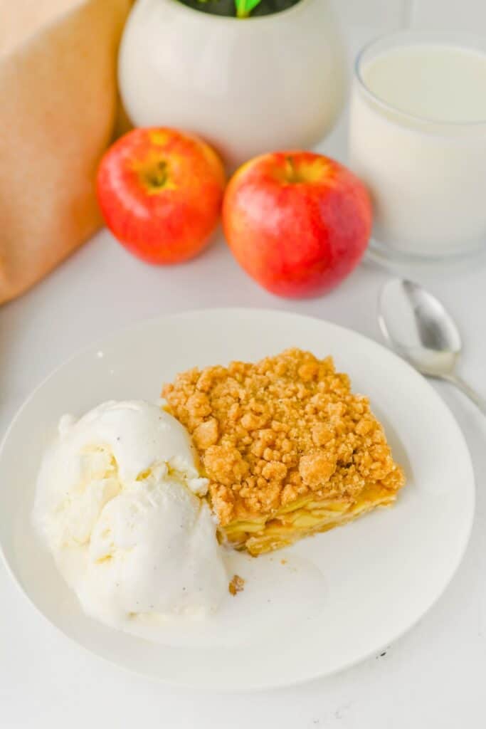 A slice of Apple Pie Bars potluck dessert with layers of cinnamon apples and a golden crumb topping, served on a white plate with two scoops of vanilla ice cream.