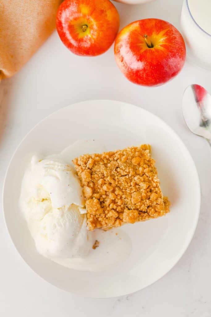 Easy apple pie bar with a golden crumb topping served beside two scoops of melting vanilla ice cream. Two red apples and a glass of milk sit above the plated dessert.