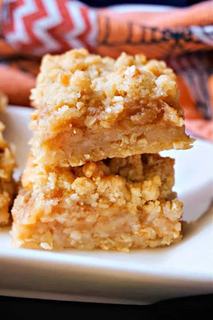 Two thick apple crumble bars are stacked on a white plate, showcasing a soft, spiced apple filling layered between a buttery, golden oat crumble crust and topping. The texture and warm cinnamon flavor make these bars a cozy, crowd-pleasing dessert.