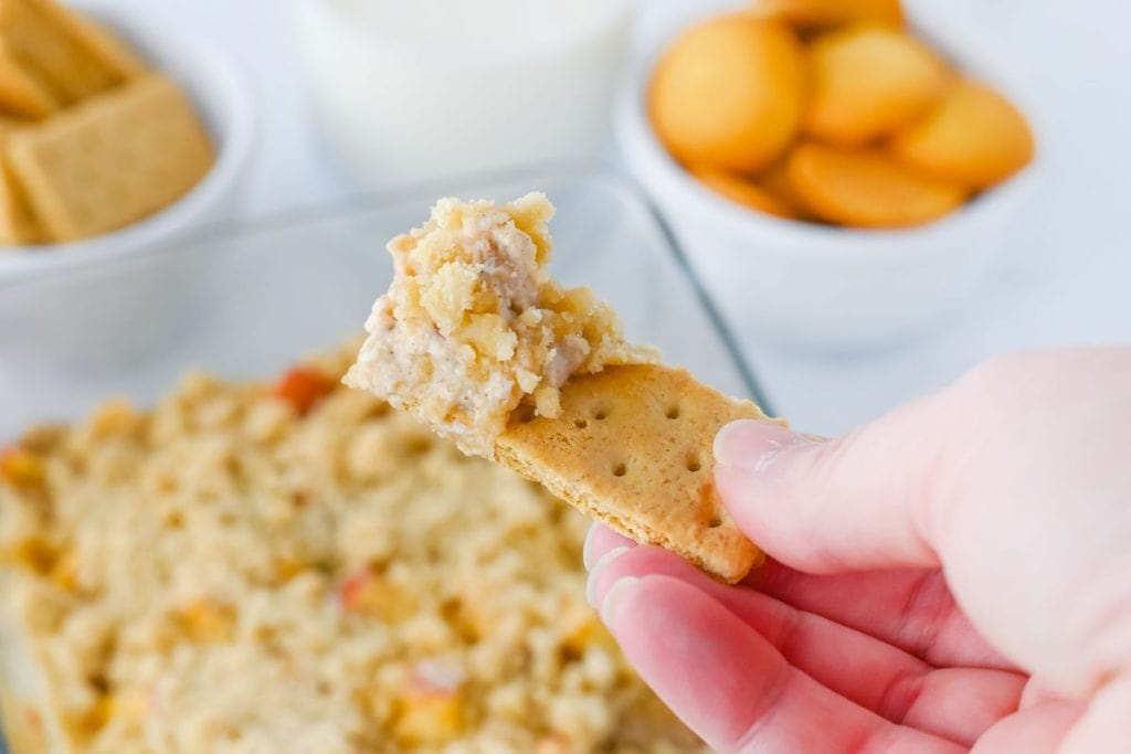 Close-up of a graham cracker holding a scoop of apple cinnamon cream cheese dip, showing the creamy, spiced filling with bits of apple and crumb topping. Blurred in the background are bowls of cookies, graham crackers, and the baking dish of dip.