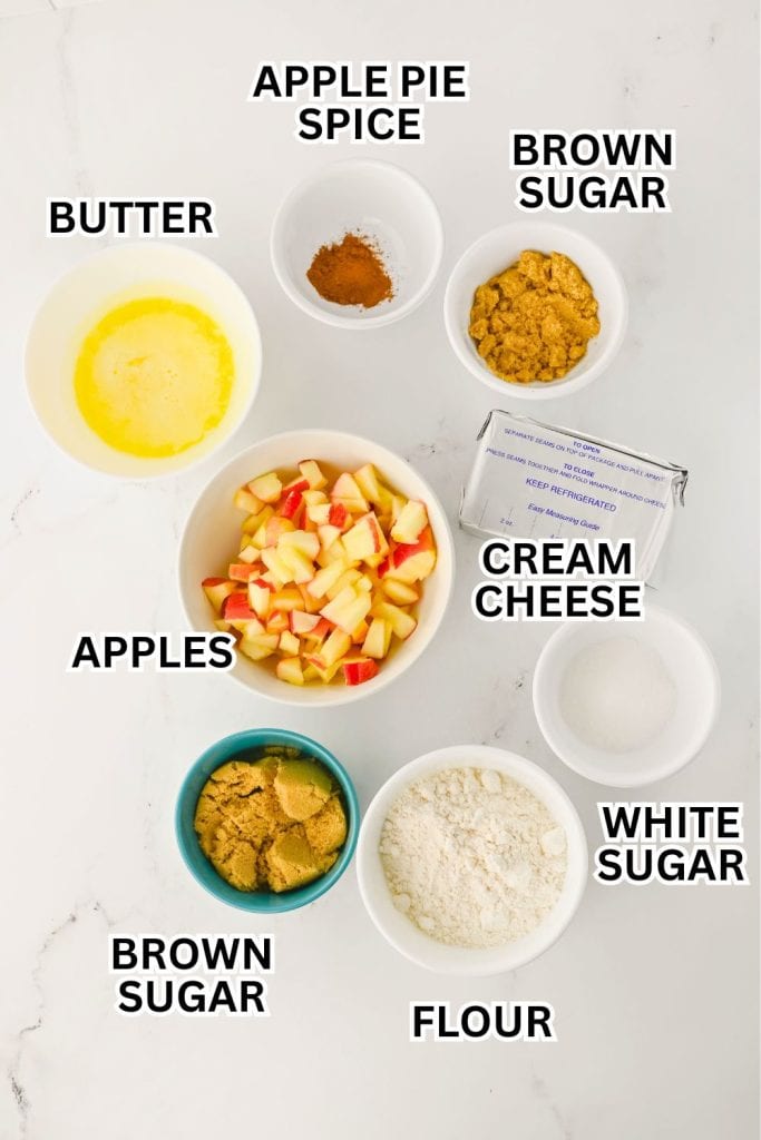 Overhead view of labeled ingredients for apple pie cheesecake dip, including diced apples, cream cheese, brown sugar, white sugar, flour, melted butter, and apple pie spice. Each ingredient is placed in a separate bowl on a white surface for easy reference.
