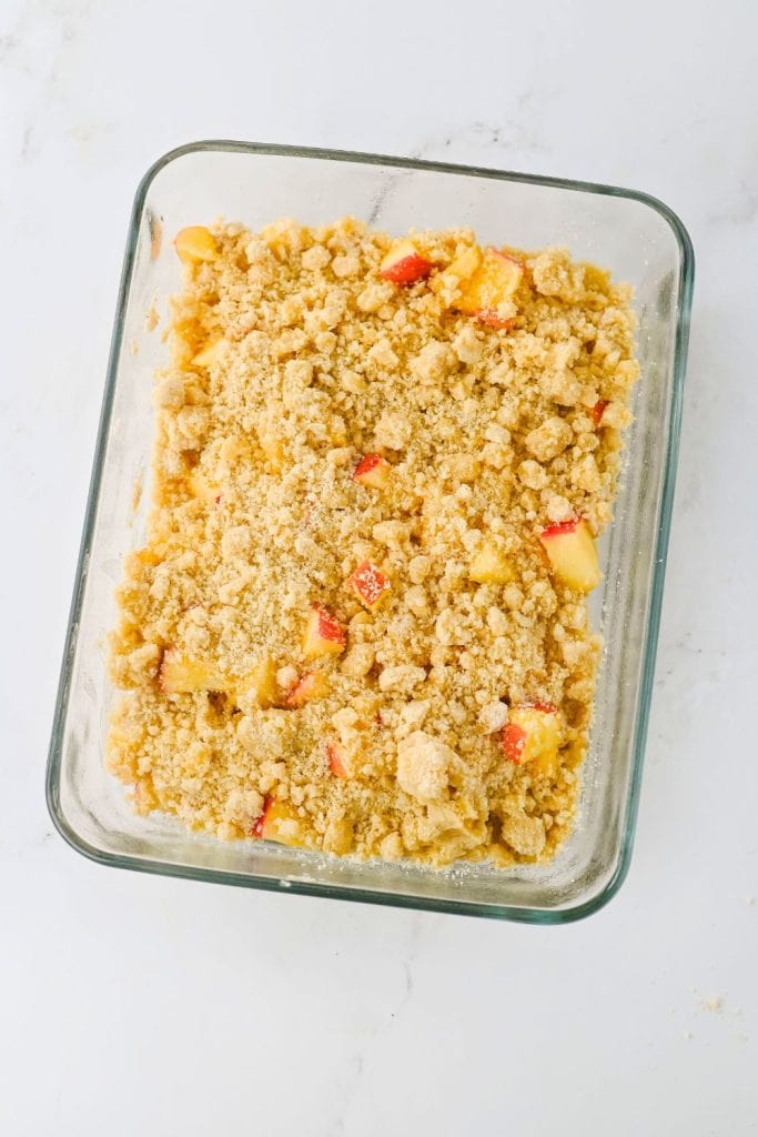 Unbaked apple cream cheese dip in a glass dish, topped with a thick layer of crumbly streusel and bits of fresh red apple. The dessert is ready to go into the oven on a clean white countertop.