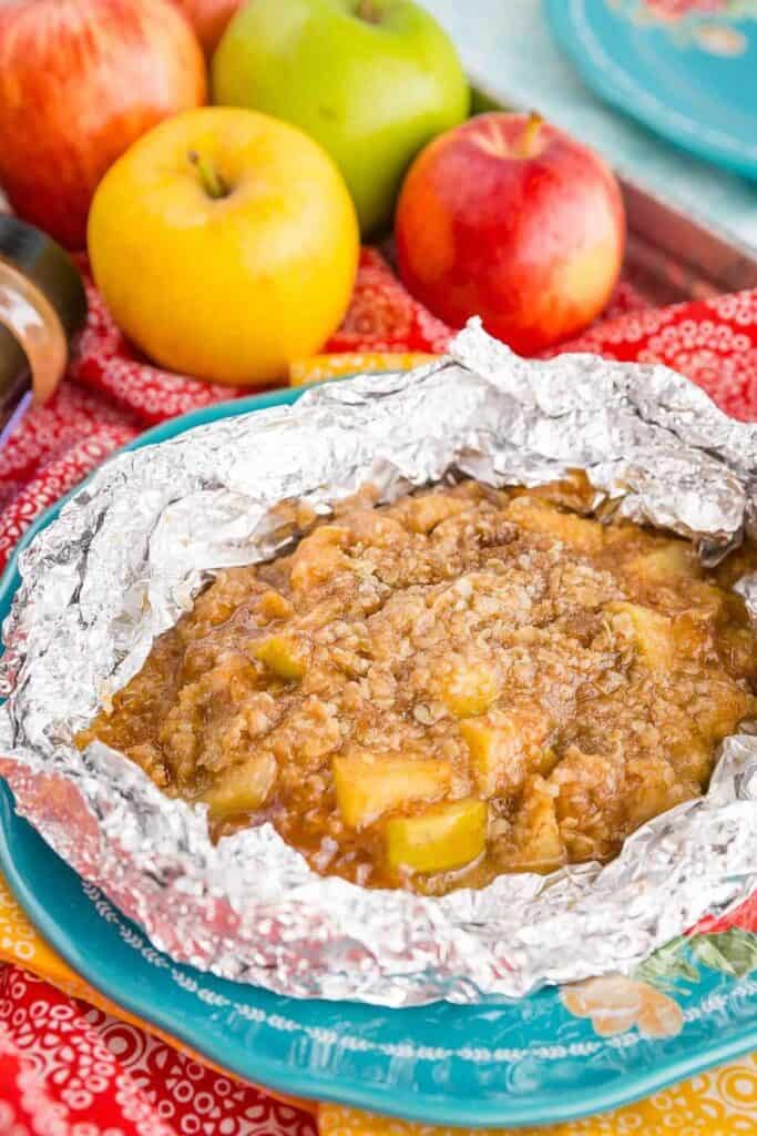 Foil packet with apple oats pie inside on a colorful tea towel with apples in the background. 