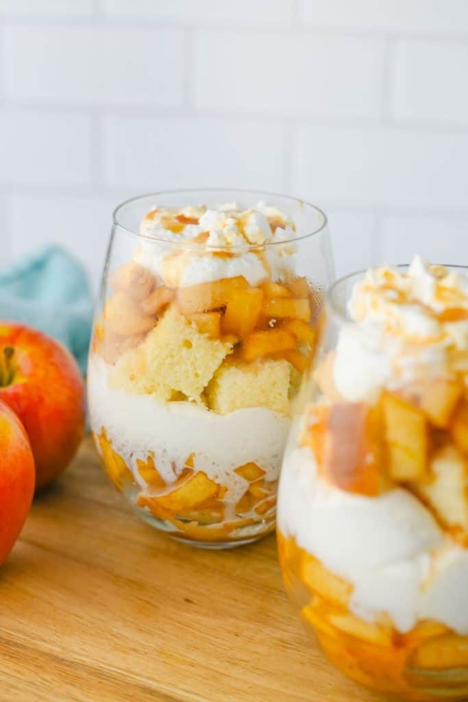 Glass cups filled with apple pie trifle are layered with whipped cream, soft cake pieces, spiced apple chunks, and topped with caramel drizzle. The dessert is served on a wooden board with whole apples and a spoon nearby, giving it a warm, homemade feel.