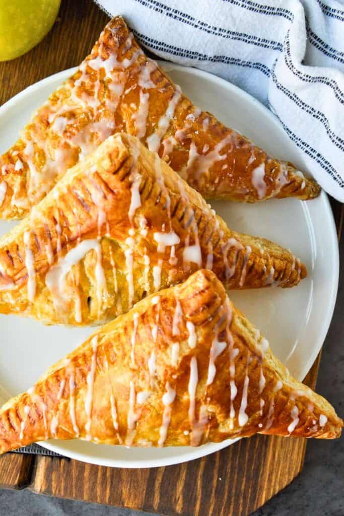 Golden brown apple turnovers are arranged on a white plate, each with a flaky, puff pastry crust and drizzled with sweet vanilla glaze.