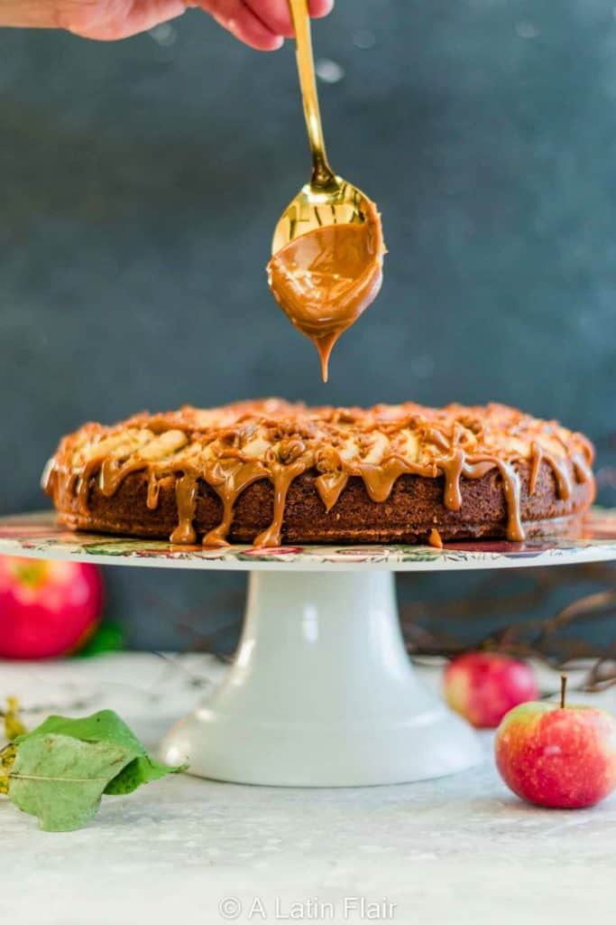 A golden apple streusel cake on a white pedestal stand, drizzled with glossy caramel sauce from a spoon held above. The crumbly topping and rich glaze highlight the cozy, spiced layers of this easy apple dessert.