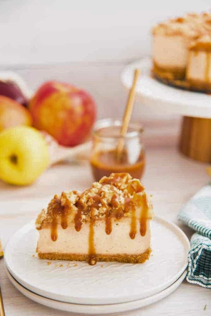 A slice of creamy cheesecake with a graham cracker crust, topped with apple crumble and drizzled with caramel sauce, served on a white plate. Fresh apples and a jar of caramel sit in the background adding a cozy autumn feel. 