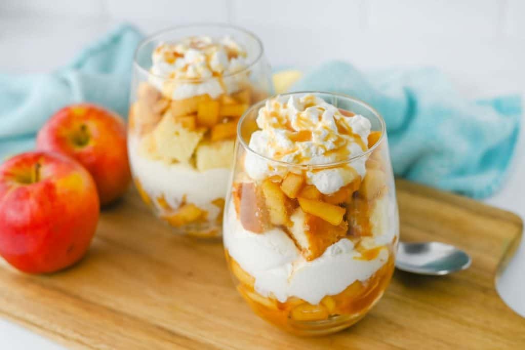 Two glass cups filled with easy holiday trifles are layered with whipped cream, spiced apple chunks, golden pound cake, and topped with a caramel drizzle. The dessert is served on a wooden board beside fresh apples and a soft blue cloth for a cozy autumn presentation.