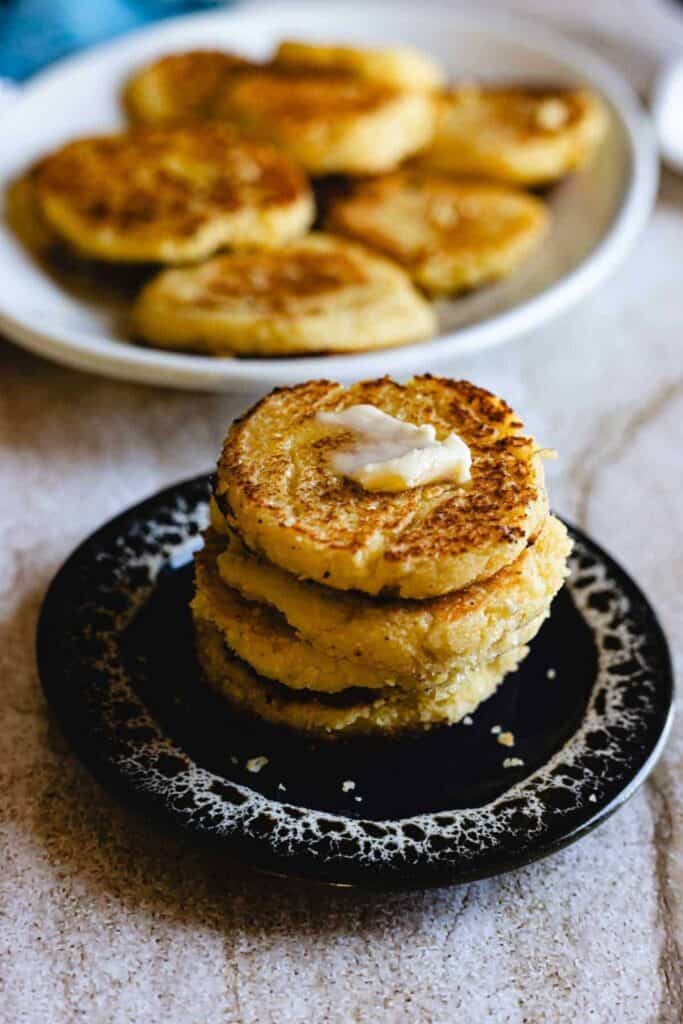 A stack of arepas de choclo that look lik corn pancakes, on a small plate topped with a pat of butter melting into the golden crispy surface. More arepas sit in the background showing their thick texture and lightly browned edges.
