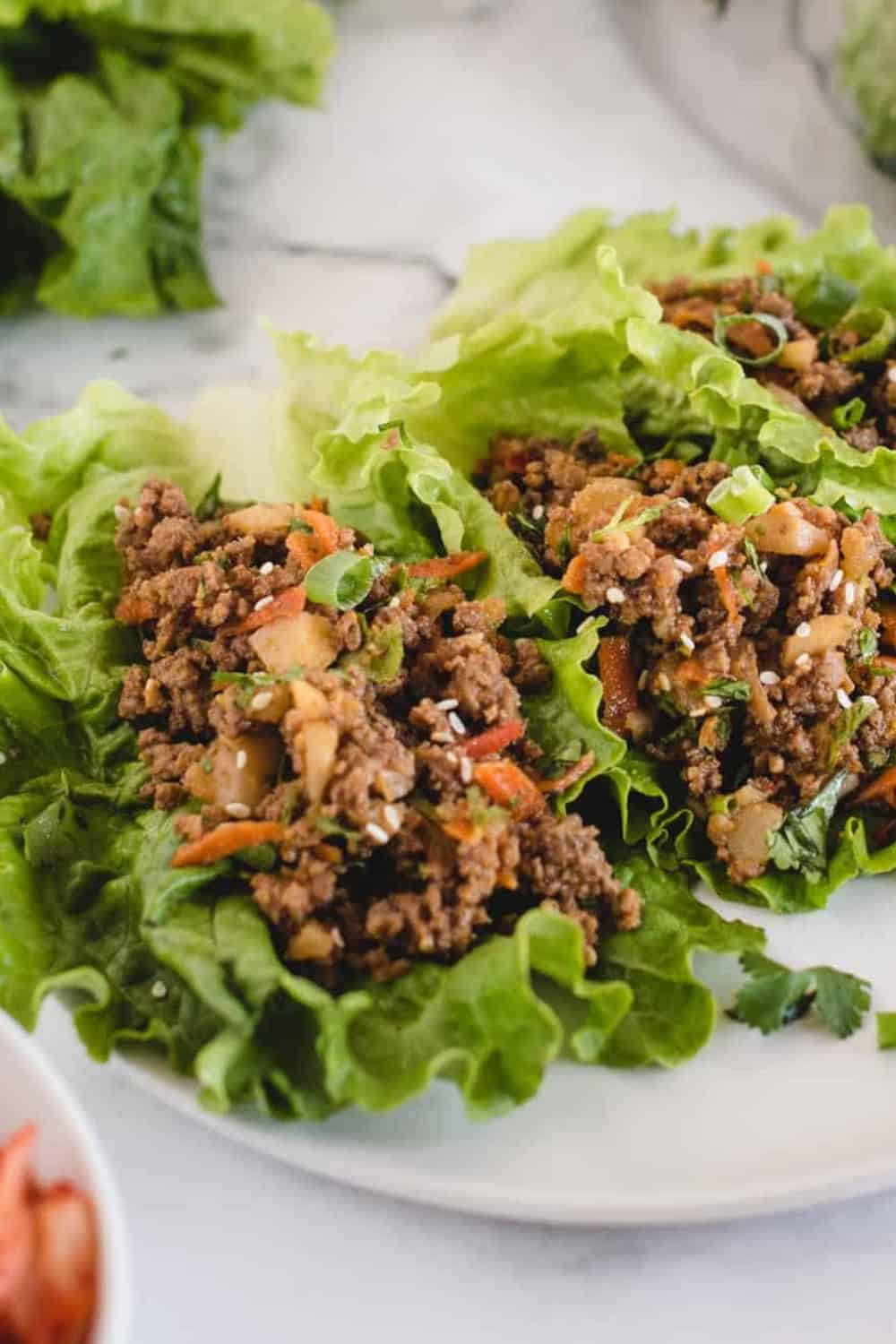 sian ground beef lettuce wraps made with seasoned ground beef, shredded carrots, water chestnuts, and fresh herbs spooned into crisp lettuce leaves. The wraps are garnished with sliced green onions and sesame seeds and served on a white plate.