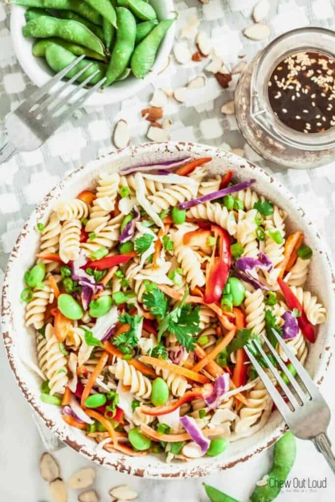 Bowl of Asian pasta salad with rotini edamame shredded carrots red bell pepper purple cabbage and green onions tossed with sesame ginger dressing and topped with cilantro and sliced almonds. A small jar of sesame dressing and a bowl of edamame sit beside the colorful cold noodle style salad.