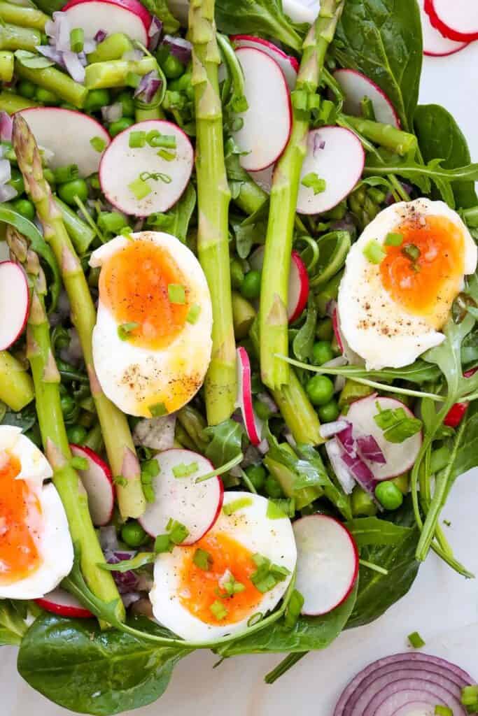 overhead view of a fresh salad featuring halved soft-boiled eggs with runny yolks, crisp asparagus spears, sliced radishes, green peas, arugula, and chopped red onion, garnished with cracked black pepper and chopped green onions.