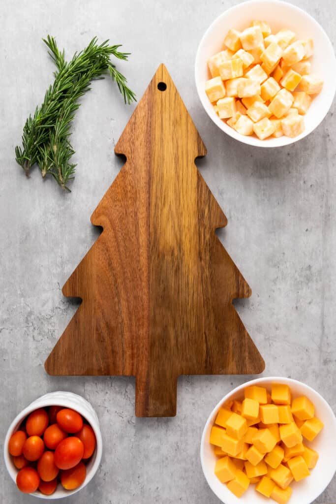 Wooden Christmas tree shaped cutting board, with small bowls of cubed cheese, cherry tomatoes and 3 sprigs of rosemary beside it. 