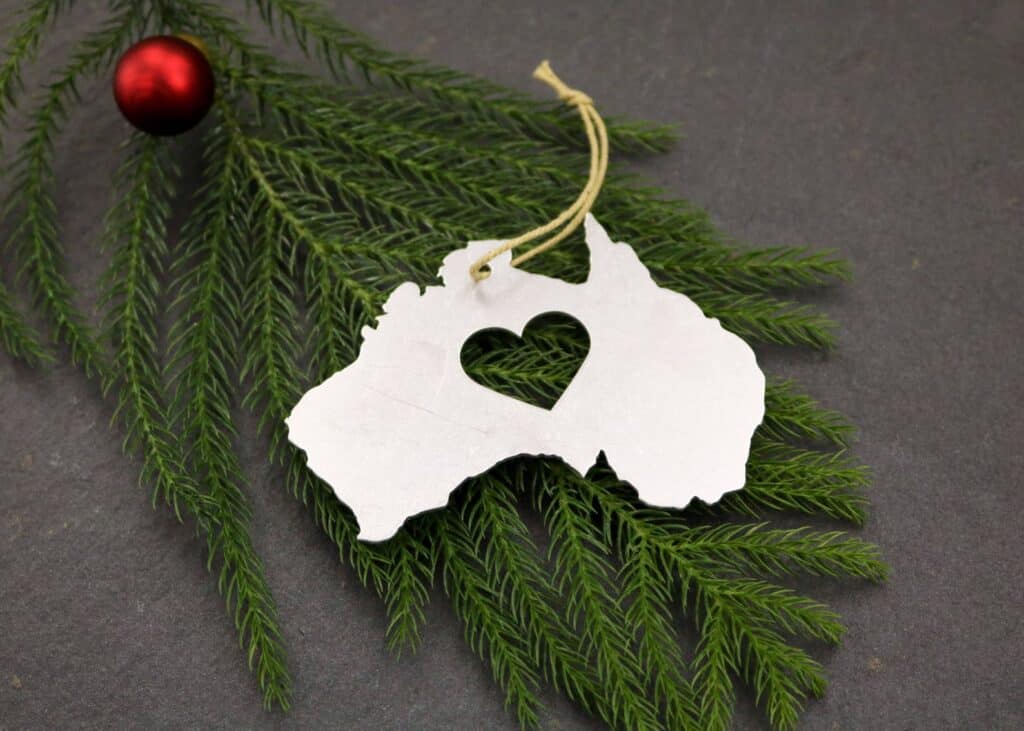 Australian Christmas Tree Ornament is the outline of the country, meade of metal, with a geart cut out in the middle. Lying on top of needle branch and Christmas Ball ornament.