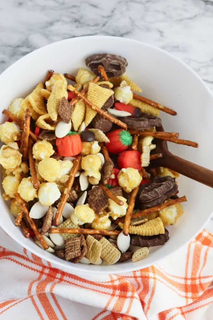 White bowl filled with autumn snack mix, featuring caramel popcorn, Chex cereal, Bugles, pretzel sticks, chocolate-covered pretzels, candy pumpkins, candy corn, pumpkin seeds, and mini peanut butter cups, with a wooden spoon resting inside and a plaid orange towel underneath.