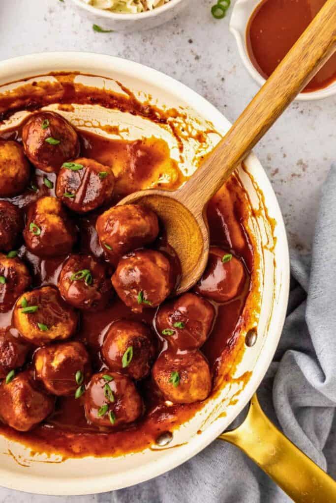 BBQ chicken meatballs simmering in a thick, glossy barbecue sauce, garnished with chopped green onions and served in a white skillet with a wooden spoon, ready for scooping and serving.