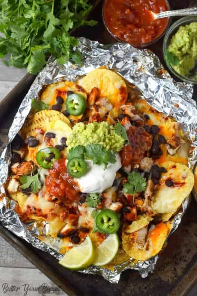 Overhead view of a foil packet with BBQ chicken nachos topped with cheese, black beans, a dollop of sour cream and guacamole on top with jalapeno slices. 