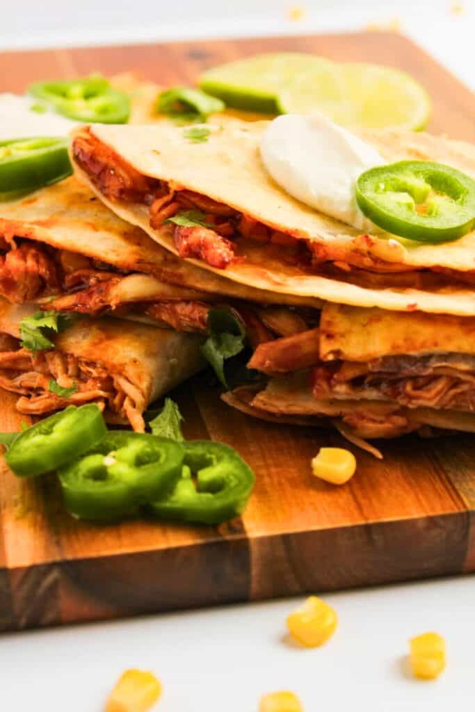 BBQ chicken quesadillas stacked on a wooden cutting board with melted cheese and shredded barbecue chicken tucked inside toasted tortillas. Sliced jalape&ntilde;os, lime wedges, cilantro, and a dollop of sour cream are served alongside the quesadillas.