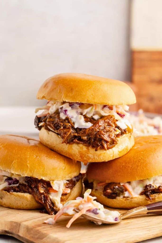 BBQ pulled pork sliders stacked on a wooden board, each filled with shredded barbecue pork and creamy coleslaw on soft golden buns. A spoonful of extra slaw rests in front for serving.