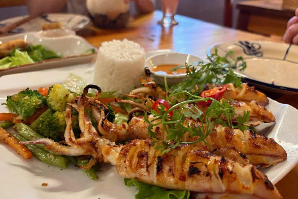 Close-up of a grilled BBQ squid dish served with steamed white rice, stir-fried vegetables, fresh herbs, and a dipping sauce. The meal is presented on a white plate at a wooden table with drinks, Spring rolls,  and other dishes in the background.