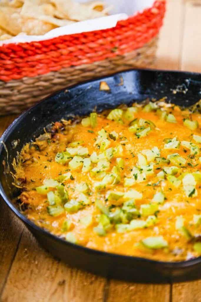 Hot Bacon Cheeseburger Dip served in a black skillet, topped with melted cheddar cheese, chopped pickles, and parsley. A basket of tortilla chips sits nearby, ready for dipping into this hearty, cheesy appetizer.