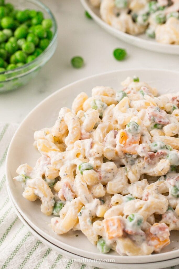 Creamy bacon ranch pasta salad served on a white plate, featuring twisted pasta, green peas, diced cheddar cheese, and crispy bacon bits coated in a ranch dressing. A bowl of fresh green peas is visible in the background on a light countertop.