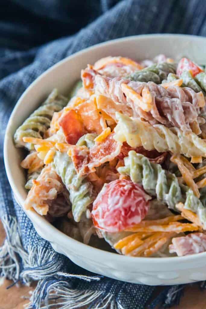 Bowl of bacon ranch pasta salad with rotini pasta crispy bacon cherry tomatoes shredded cheese and tender chicken tossed in a creamy ranch dressing. The pasta salad is served in a white bowl on a wooden table with a blue cloth underneath.