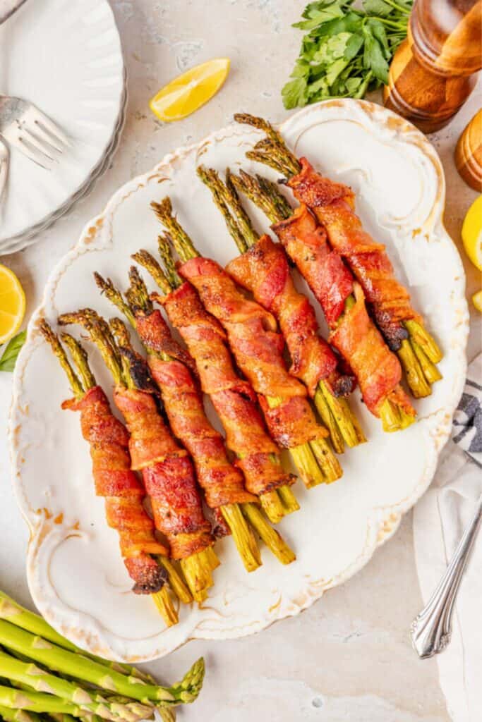 Bundles of roasted asparagus spears wrapped in crispy bacon are arranged on an ornate white serving platter. Lemon wedges, fresh parsley, and additional asparagus surround the platter for a colorful presentation.
