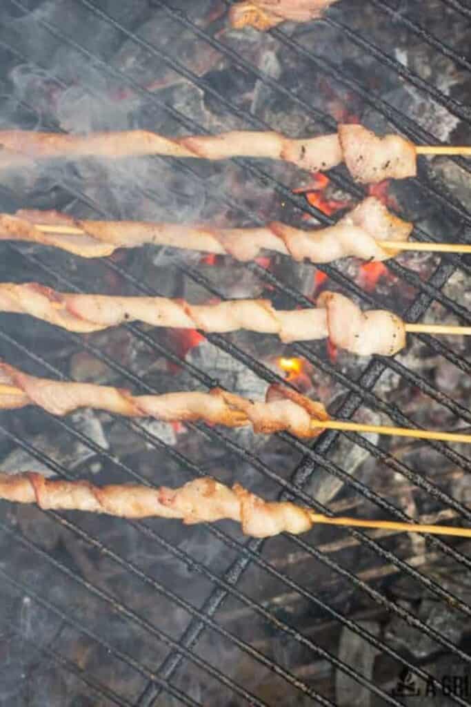 Wooden skewers wrapped in bacon cooking over a campfire grill.