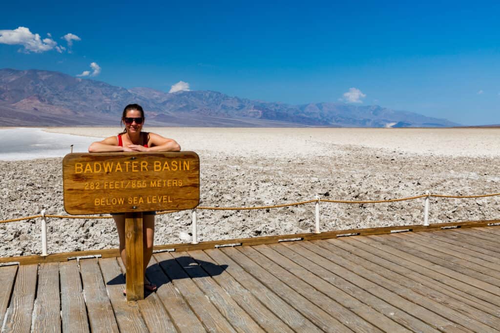 Woman in tank top standing behind the Badwater Basin wooden sign, with the badlands behind and mountains in the distance.