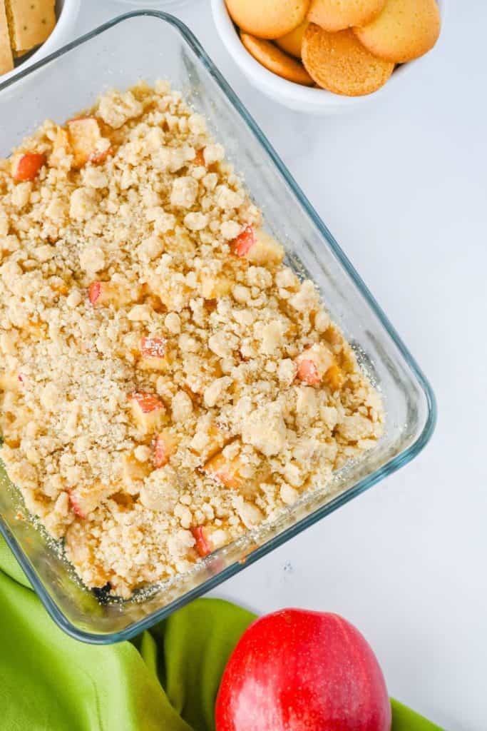 Glass dish filled with freshly baked apple dip, topped with golden brown streusel and chunks of red apples. Surrounded by vanilla wafers, graham crackers, and a red apple on a green cloth for a cozy serving setup.