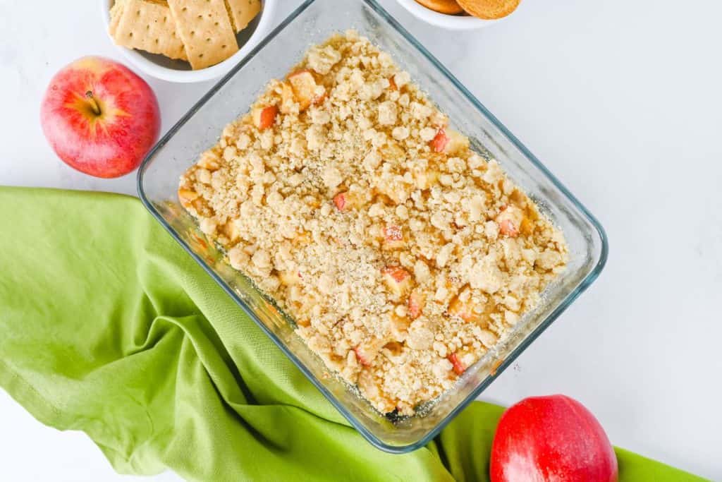 glass baking dish filled with baked apple pie dip, topped with a golden, crumbly streusel. Surrounding the dish are fresh red apples, graham crackers, and cookies, with a green cloth adding contrast.