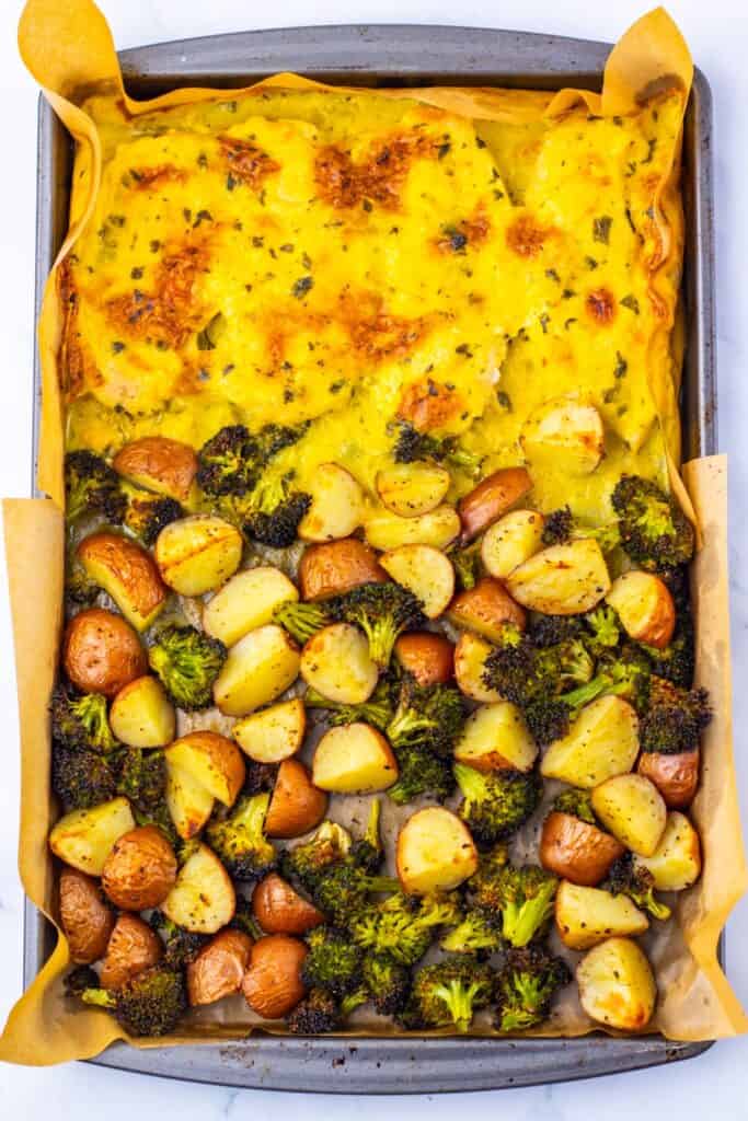 Baked chicken breast, potatoes, and broccoli sheet pan recipe fresh from the oven, with golden, cheesy chicken on one side and roasted baby potatoes and broccoli on the other. Everything is cooked on a parchment-lined baking sheet for an easy, one-pan meal.