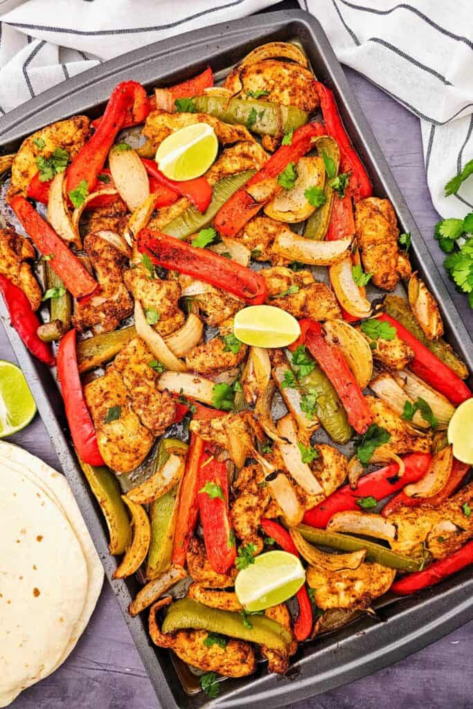 Baked chicken fajitas oven style with seasoned chicken strips roasted on a sheet pan with red and green bell peppers and sliced onions. The pan is garnished with fresh cilantro and lime wedges and served with warm tortillas for an easy family meal.
