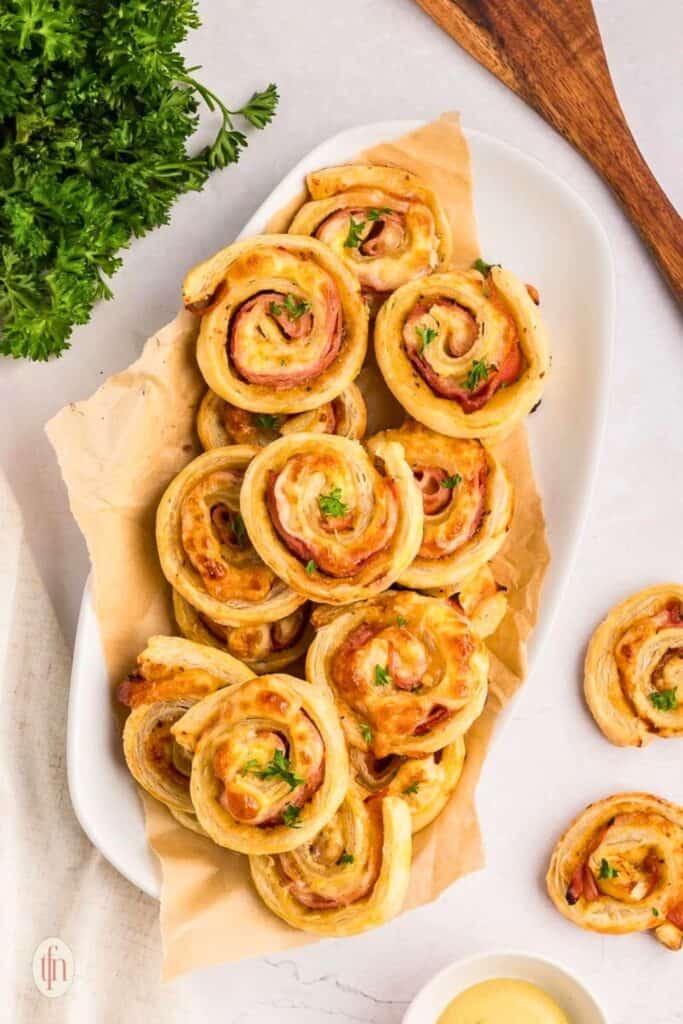 A platter of golden-brown baked ham and cheese pinwheels made with puff pastry, swirled with melted cheese and thinly sliced ham, garnished with fresh parsley and served on parchment paper with a side of dipping sauce.