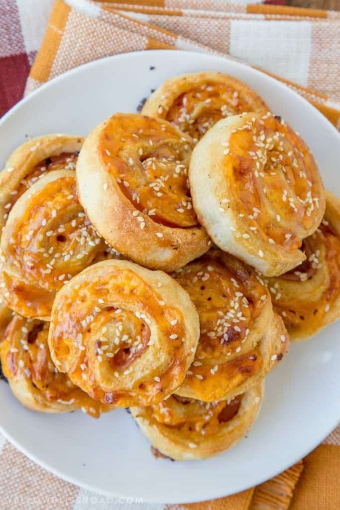 Golden baked ham and cheese roll ups topped with melted cheddar and sprinkled with sesame seeds, stacked on a white plate. These savory party finger foods are swirled in a pinwheel shape with visible layers of ham and cheese.