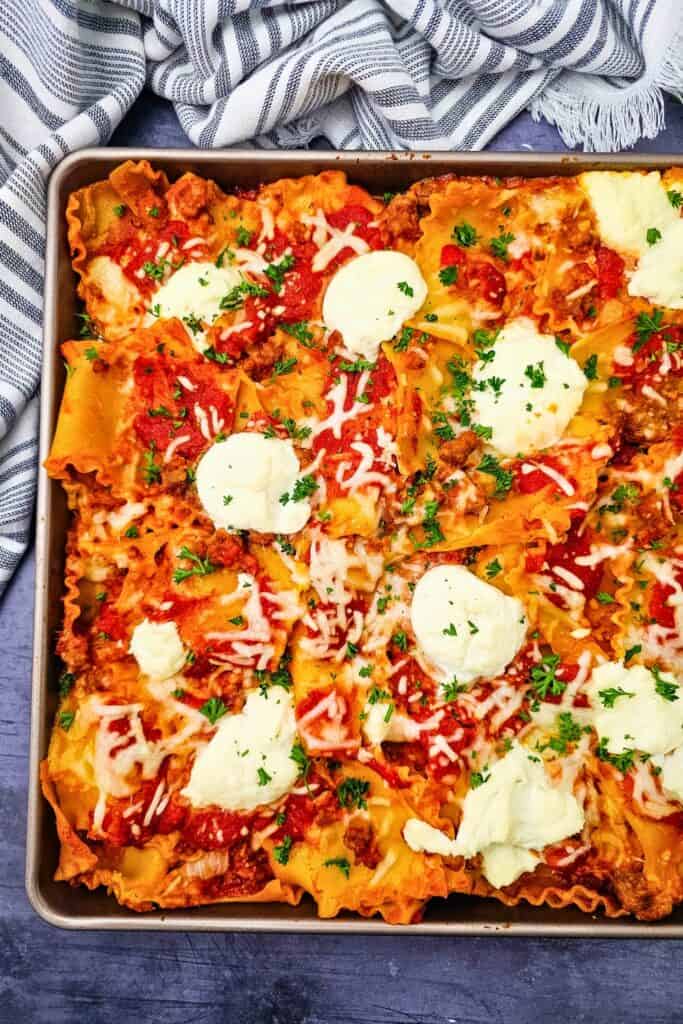 Baked sheet pan lasagna with ruffled noodles covered in Italian sausage meat sauce and melted mozzarella. Creamy ricotta and fresh parsley are scattered across the golden baked pasta.