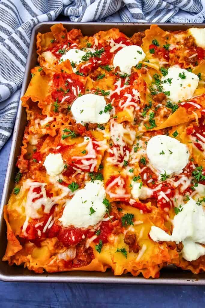 Side view of a sheet pan dinner with ruffled lasagna noodles covered in tomato meat sauce and melted mozzarella. Generous dollops of ricotta and chopped fresh parsley top the rustic lasagna, beside a striped cloth.