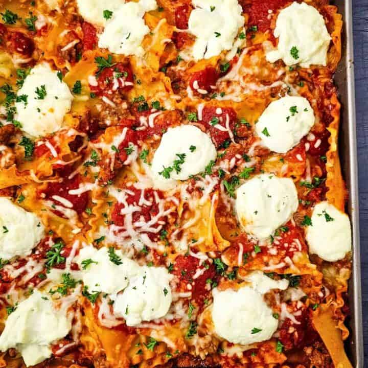 Lazy lasagna spread across a sheet pan with ruffled noodles covered in tomato meat sauce and melted cheese. Large dollops of ricotta and fresh parsley top this easy sheet pan dinner.