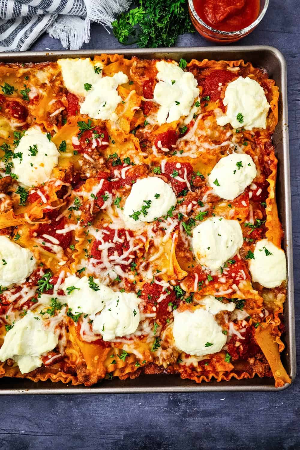 Lazy lasagna spread across a sheet pan with ruffled noodles covered in tomato meat sauce and melted cheese. Large dollops of ricotta and fresh parsley top this easy sheet pan dinner.