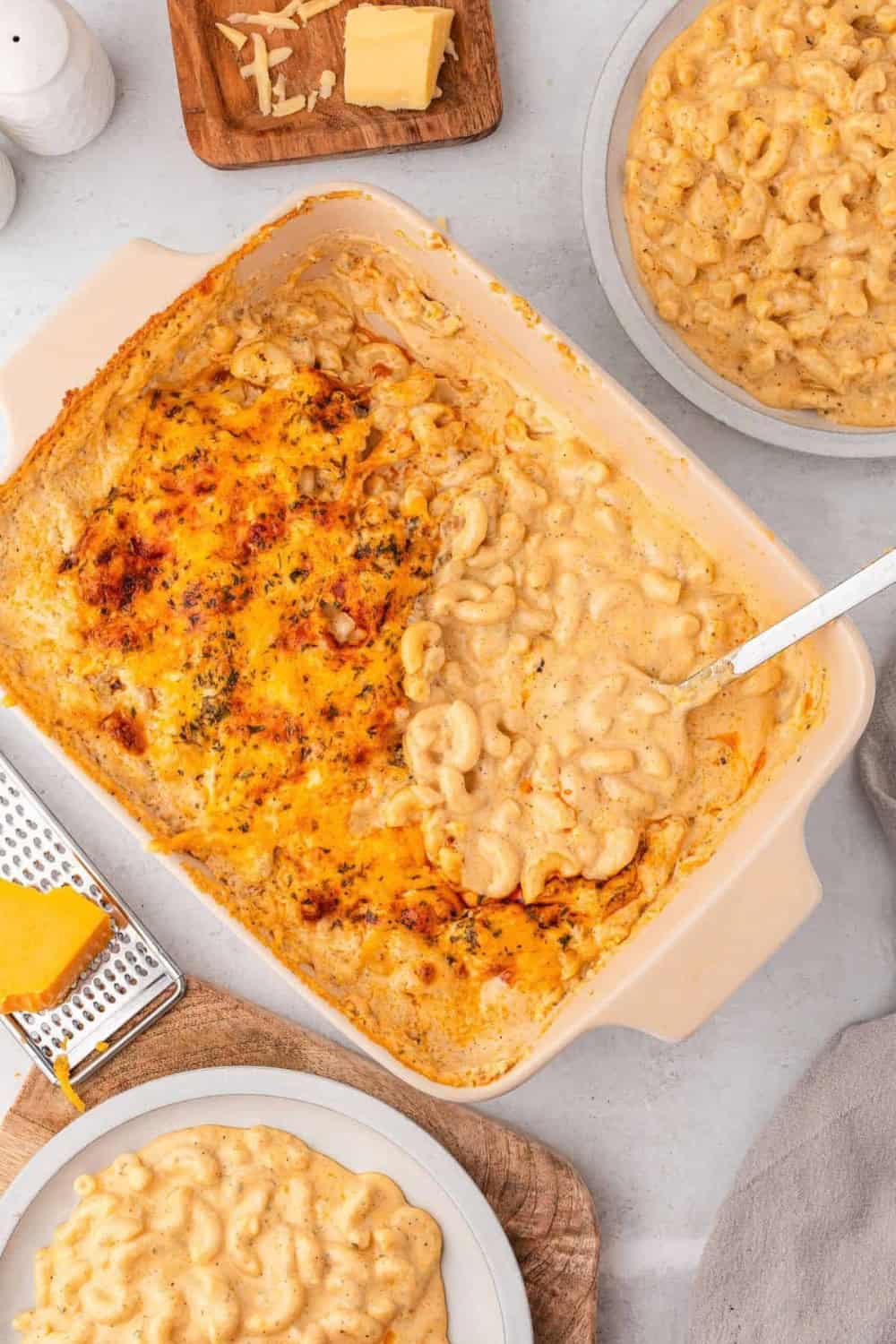 An overhead view of creamy baked macaroni and cheese in a casserole dish with tender elbow pasta in a rich cheese sauce and a golden, browned cheese topping, with portions served on plates alongside shredded and block cheese.