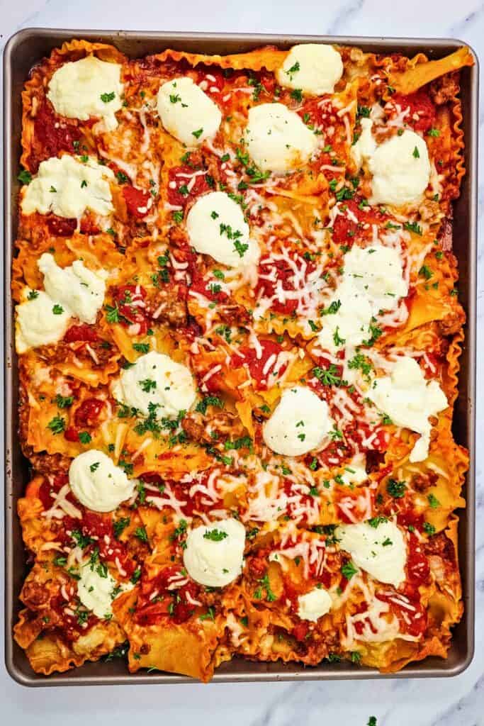 Overhead view of a baked one pan layerless lasagna with ruffled noodles covered in Italian sausage tomato sauce and melted mozzarella. Large dollops of creamy ricotta and fresh parsley are scattered across the top.
