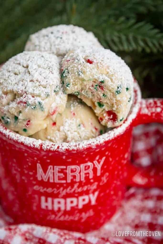 Soft, round Christmas snowball cookies with red and green sprinkles are dusted with powdered sugar and stacked in a red mug that reads “Merry everything & Happy always.”  The festive cookies sit atop a red gingham cloth with pine branches in the background ready for a cookie exchange party.