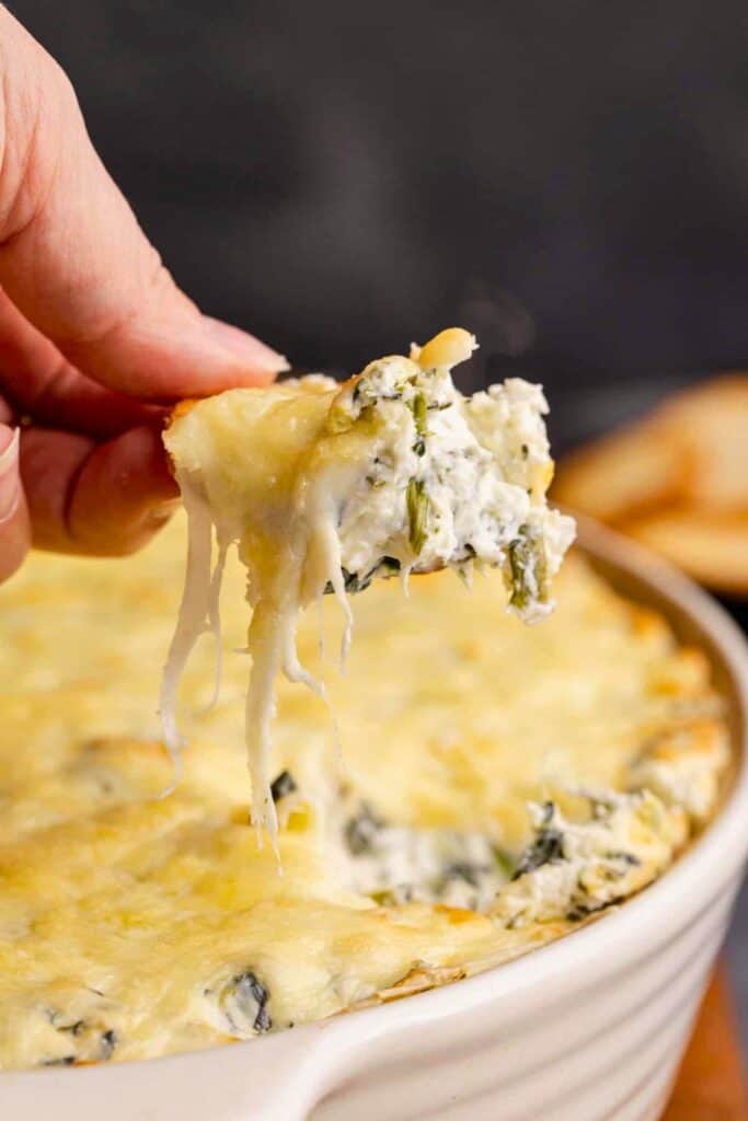 A hand lifts a tortilla chip loaded with creamy baked spinach artichoke dip from a serving dish. Long strands of melted cheese stretch from the hot golden dip to the chip.