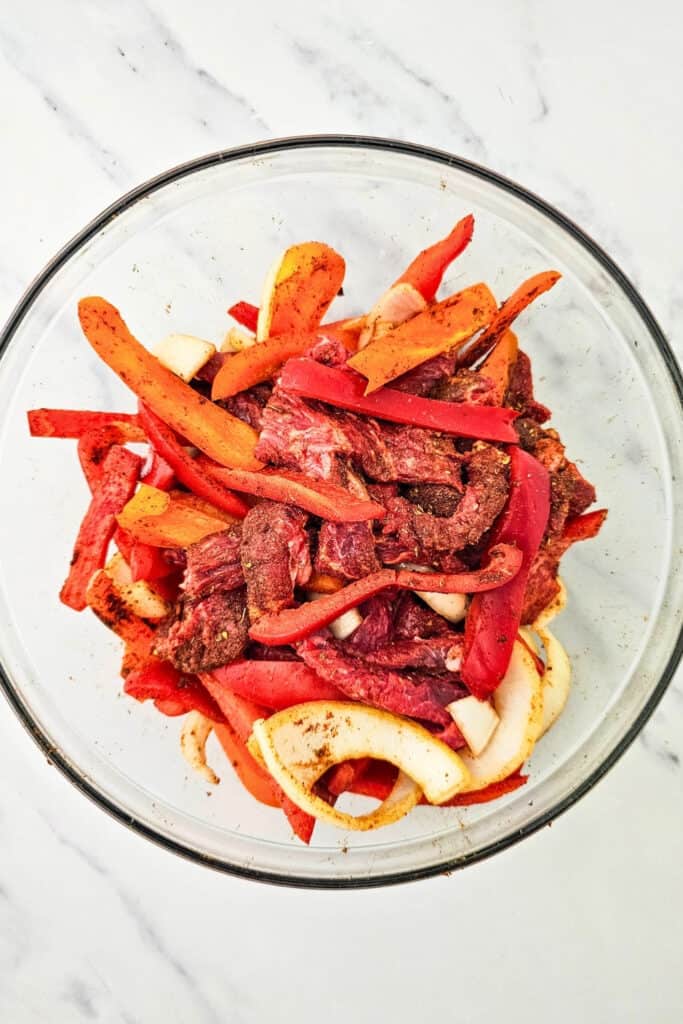 Raw steak strips are tossed with sliced red and orange bell peppers and white onions in a large glass bowl. The steak and vegetables are evenly coated with fajita seasoning and ready to be spread on a sheet pan for baking.