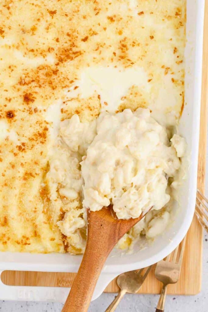 Creamy baked white cheddar mac and cheese in a white casserole dish, with a golden-brown crust on top. A wooden spoon scoops out a serving, showing the rich, cheesy pasta underneath.