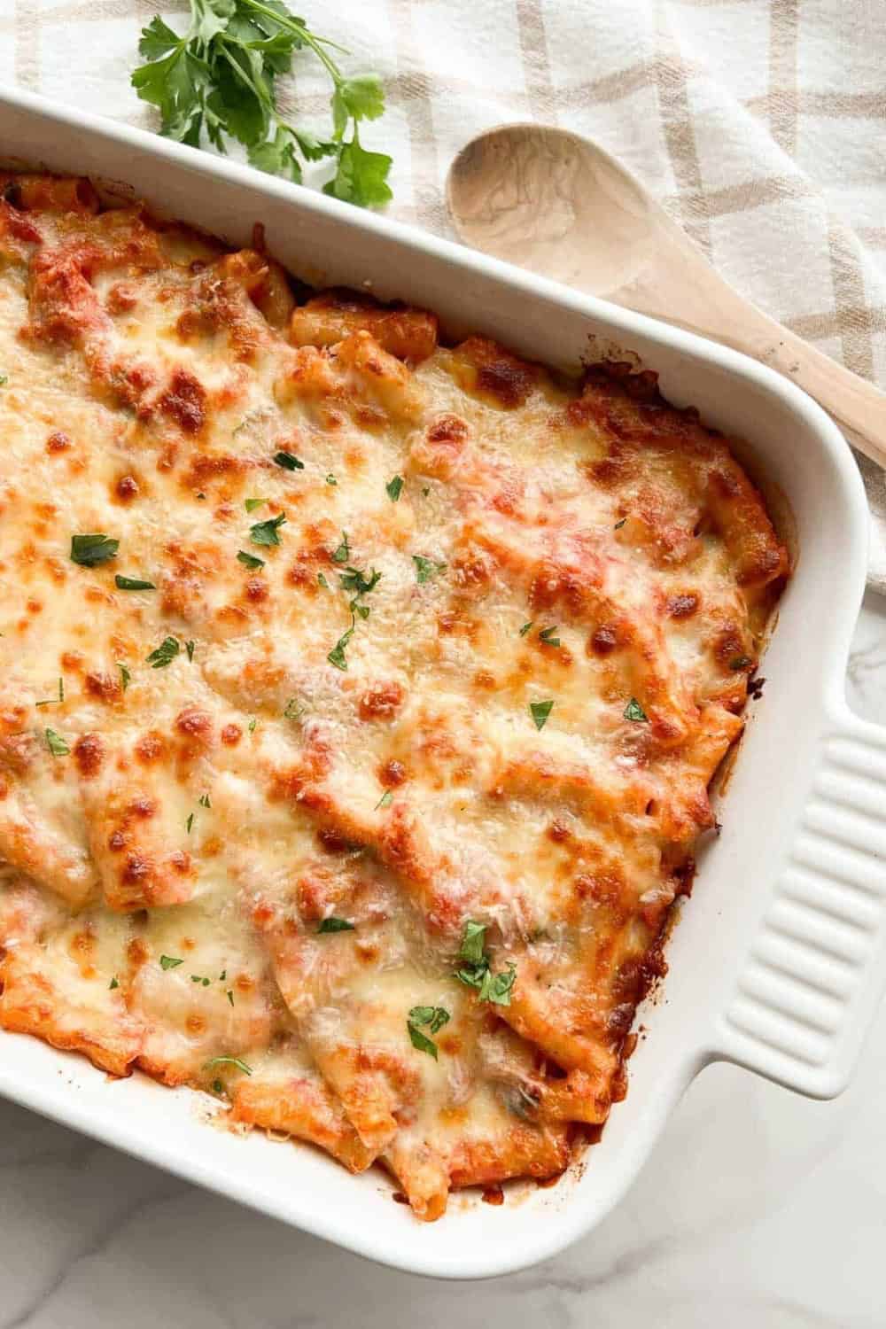 A white baking dish filled with Baked Ziti with Sausage, layered with pasta and tomato sauce under a generous layer of melted, golden-brown cheese and finished with chopped fresh parsley.