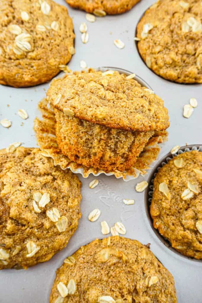 Bakery style oatmeal muffins with domed tops and oats sprinkled over a golden crust baked in a muffin tin. One muffin is lifted to show a soft hearty interior while others sit around it with a rustic texture.