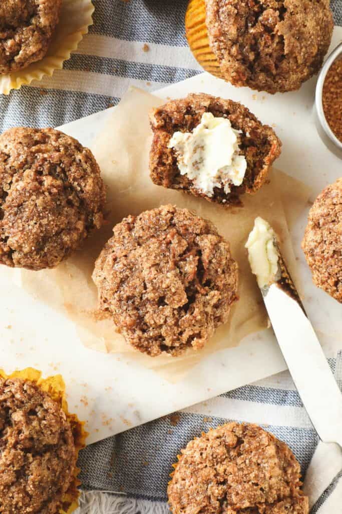 Overhead view of moist banana carrot muffins with a golden, crunchy sugar topping, one sliced open and slathered with melting butter. A butter knife and scattered raw sugar granules add a cozy, homemade touch