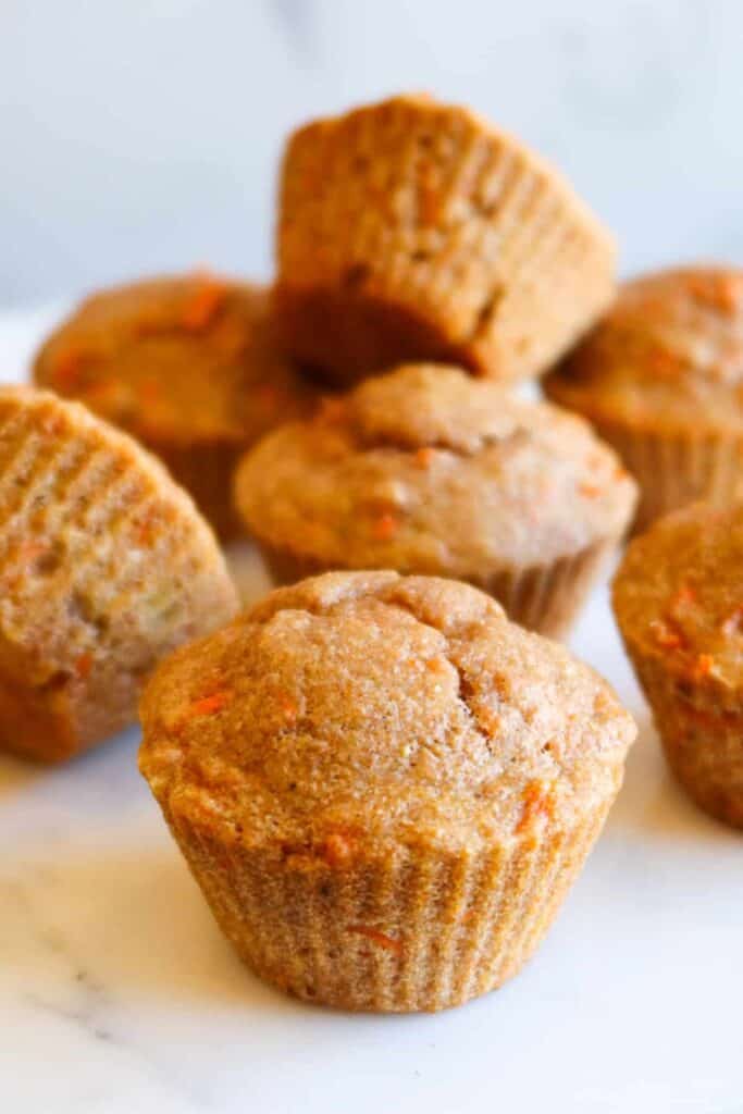 Golden brown banana carrot muffins arranged on a white surface, with one muffin in sharp focus and several stacked softly in the background. The muffins have a slightly domed top and visible flecks of shredded carrot, highlighting their moist texture and wholesome ingredients.